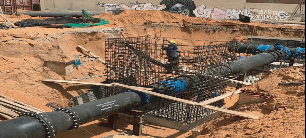 Ouvrier dans une cage d'armatures autour d'une conduite en fonte noire et de vannes bleues, au fond d'une fouille.