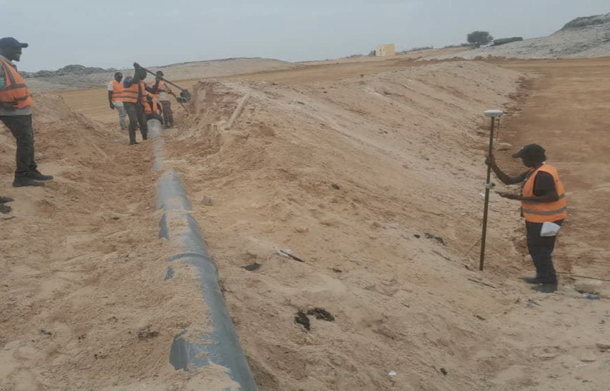 Équipe en gilets orange posant une conduite dans une tranchée, pendant qu'un topographe relève les cotes au GPS.
